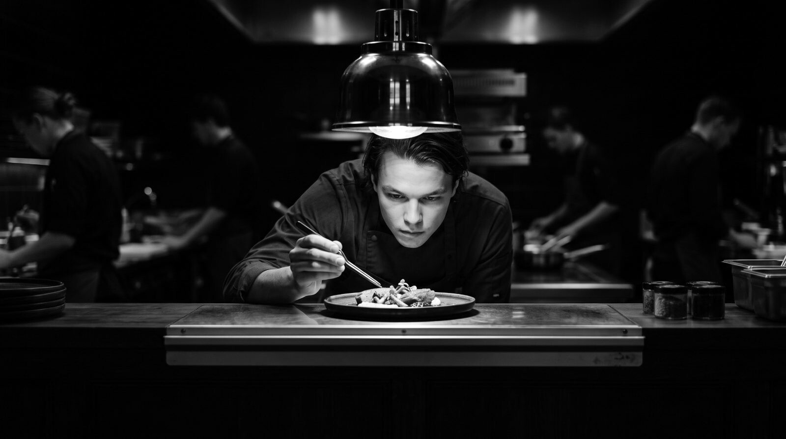 The pass, head chef inspecting a plate under a single warm lamp, kitchen dark behind