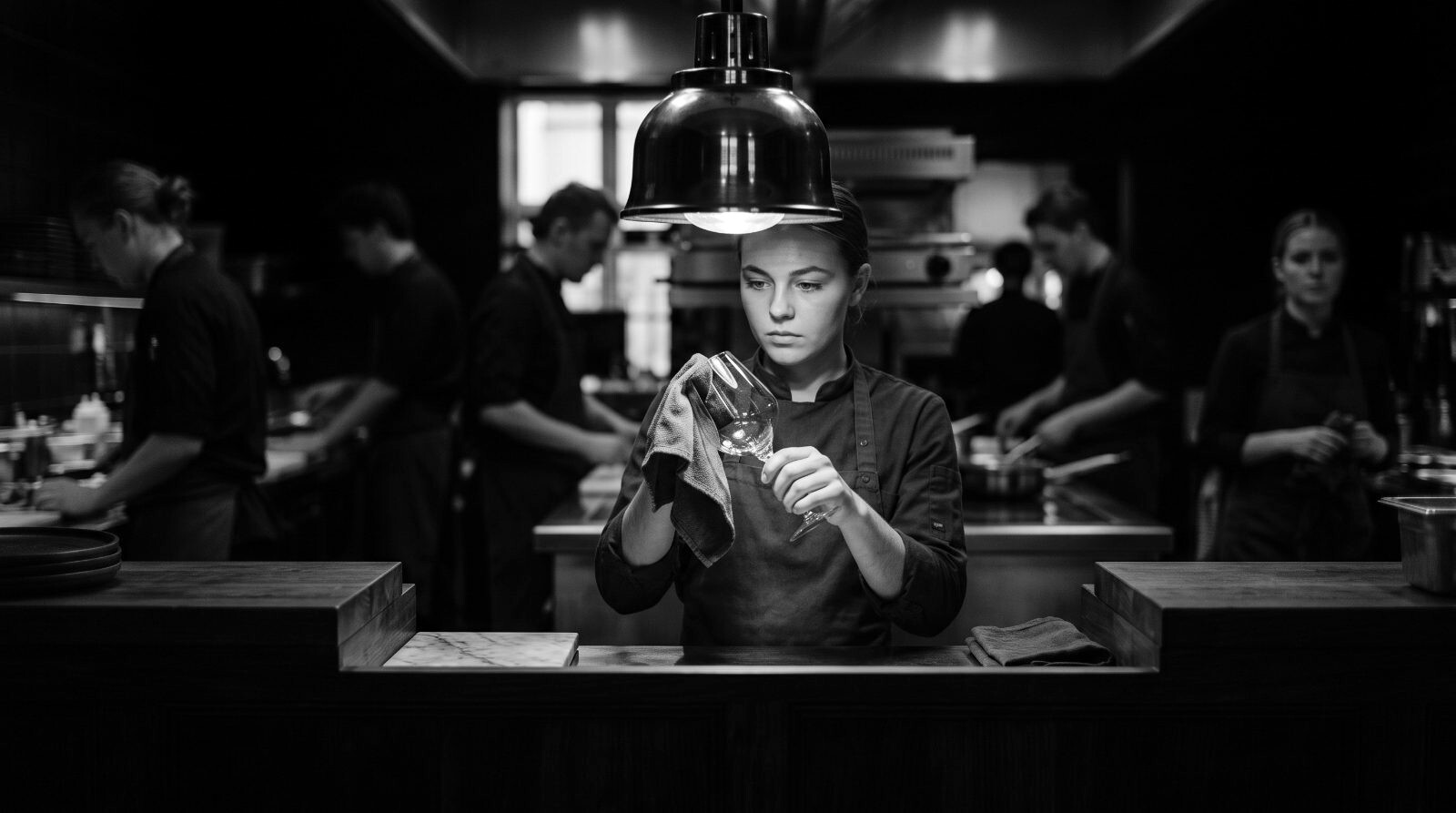 Young server polishing a wine glass to the light, brigade at work blurred behind