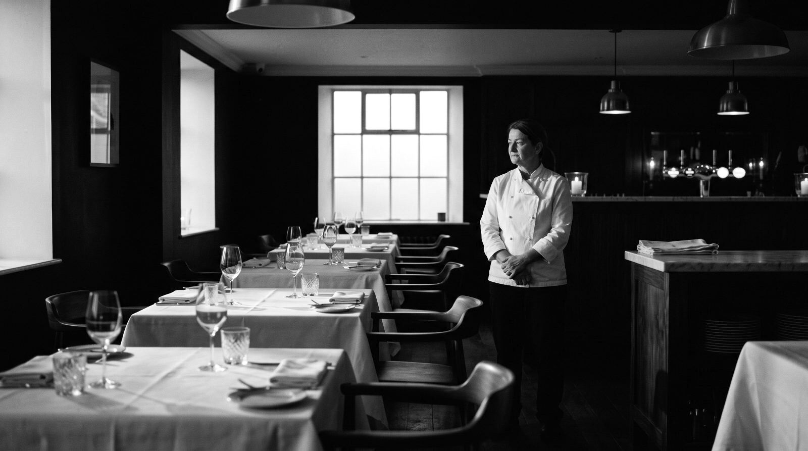 Empty dining room at dawn, tables set with impossible precision; one person surveying the room